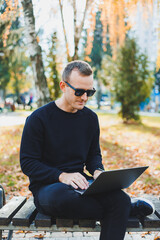 Obraz premium A young man sits in the park on a bench and works on a laptop. A warm autumn day. Remote work