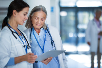 Older doctor giving advise to his younger colleague, discussing at hospital corridor. Health care concept.