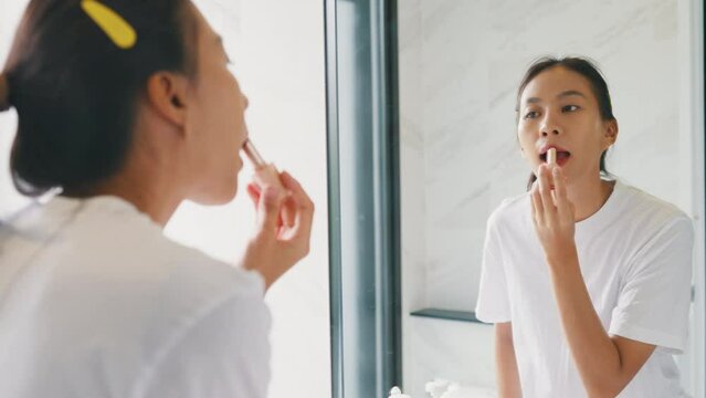 Young Asian happy woman make up looking at her reflection in mirror apply lip balm on her lips in bathroom. Lifestyle and people, Lips Protection, Natural Skin Care Cosmetics, Morning routine concept.