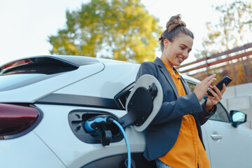 Young woman with smartphone waiting while her electric car charging in home charging station, sustainable and economic transportation concept. - obrazy, fototapety, plakaty