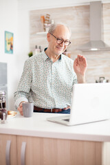Old man saying hello during video call with family using notebook in kitchen while holding a cup of coffee.