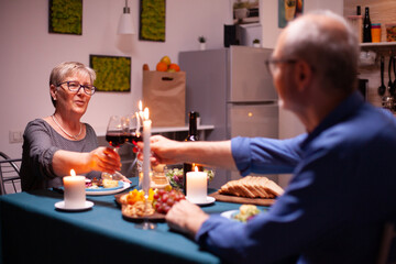 Senior couple clinking glasses with red wine during evening in kitchen. Elderly couple sitting at the table in kitchen, talking, enjoying the meal, celebrating their anniversary in the dining room.