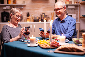 Fototapeta na wymiar Happy old couple smiling using phone in kitchen having romantic dinner. Sitting at the table in the dining room , browsing, searching, using phone, internet, celebrating their anniversary