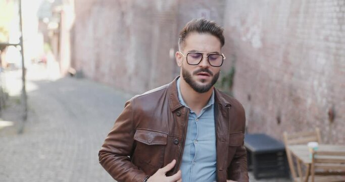 project video of sexy stylish bearded man outside in an old city walking and looking around, adjusting jacket and glasses 