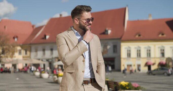 project video of attractive man with hand in pocket, touching beard and thinking while looking to side, adjusting suit and glasses and being confident outside in an old town from Romania