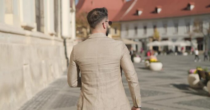 behind view of cool elegant man with fashion hairstyle walking outdoor, turning, rubbing palms and fixing suit and walking 