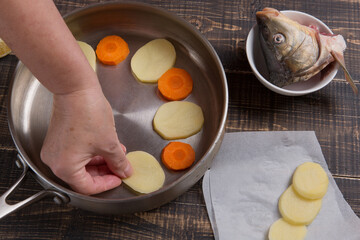 The hand of the cook puts chopped vegetables into the pan, the head of the fish lies nearby,...