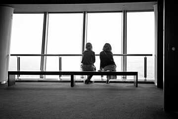 Black and white shot of two girls sitting on the bench facing the glass windows of the room