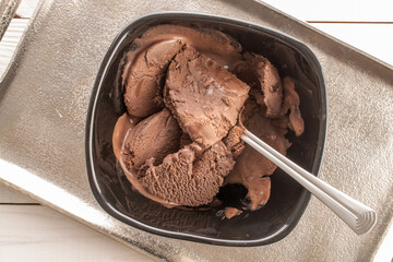 Dark brown chocolate ice cream in a black ceramic bowl with a metal spoon on a metal tray, close-up, top view.