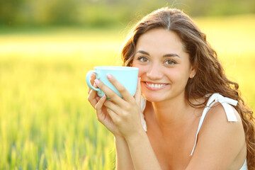 Happy woman holding coffee cup looking at camera in a field