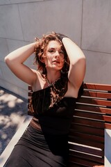 Fototapeta premium Portrait of a woman on the street. An attractive woman in a black dress is sitting on a bench outside.