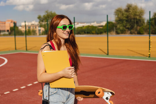 Smiling young woman walking with skateboard and file at sports court