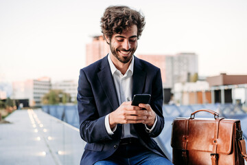 Businessman using smart phone sitting by briefcase on railing