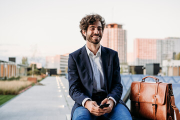 Happy businessman sitting by briefcase on railing