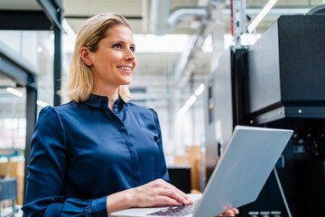 Happy businesswoman wearing blue shirt holding laptop in factory