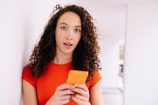 Surprised young woman with smart phone leaning on wall