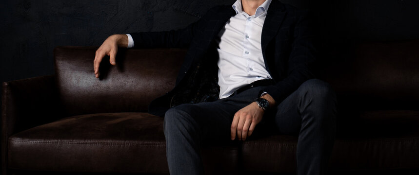 An unrecognizable man in stylish clothes sits relaxed on the sofa in the dark.