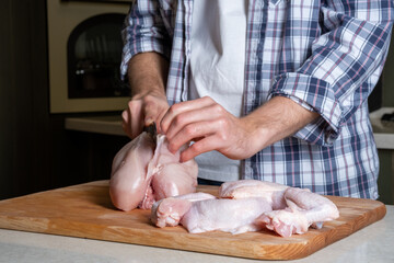 A male cook cuts fresh homemade chicken on a wooden board. Butchering the chicken in part breast, wings, legs.