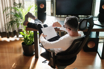 Handsome middle age man reading book at home by his computer desk near the window