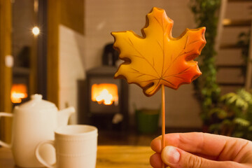 Gingerbread cookie in shape of autumnal orange maple leaf in woman's hand against burning fire in fireplace in cozy comfort home in the evening. Tea cup, teapot on a wooden table in dark room.