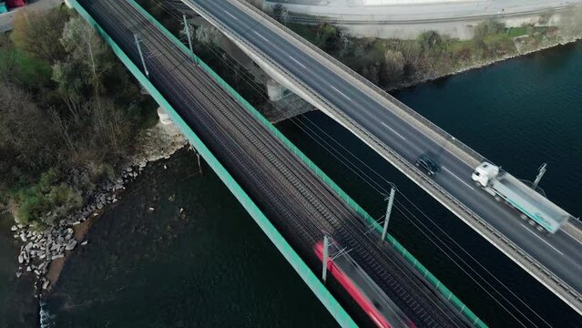 A Passenger train crossing a river via a railroad bridge located next to a highway where trucks drive next to it. Filmed with a drone slowly turning and panning.