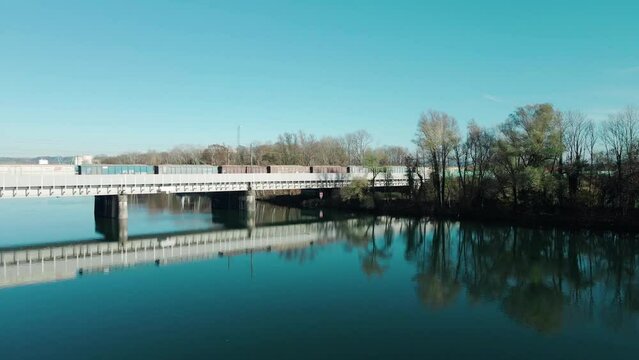 A long freight train carrying potatoes driving over a beautiful river in Austria via a railway bridge. Filmed with a drone slowly moving toward the bridge close over the water