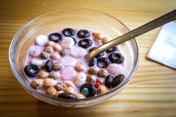 Dry breakfast in glass round plate bowl on a wooden table in cafe. Delicious cornflakes drenched in pink strawberry milk. Healthy breakfast. Food for kids. Different type cereals, multicolored balls.