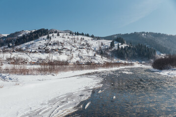 Winter, frosty day in a village located among the snow-capped mountains on the banks of the river.
