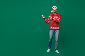 Full body merry young man 20s wear red warm knitted sweater Santa hat posing point index finger aside on workspace area isolated on plain dark green background studio. New Year 2023 holiday concept.