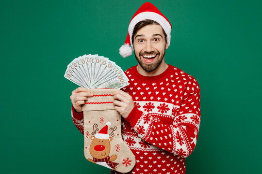 Merry young man wear red knitted sweater Santa hat posing hold in hand stocking sock fan of cash money in dollar banknotes isolated on plain dark green background studio New Year 2023 holiday concept