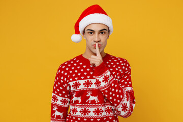 Merry young man wear knitted christmas sweater Santa hat posing say hush be quiet with finger on...