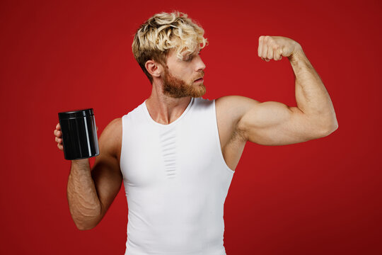Young strong sporty toned sportsman man wear white clothes spend time in home gym hold bottle of sport supplement protein show muscles isolated on plain red background. Workout sport fit body concept.