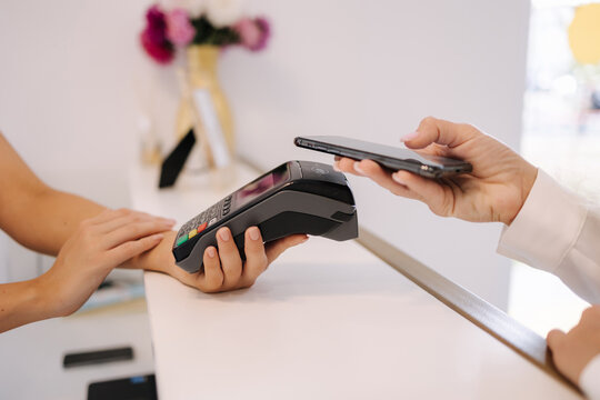 Female customer pay for procedures in beauty studio. Close-up of woman pay using phone and put to terminal. Cashier hand holding credit card reader machine while client holding phone for NFC payment