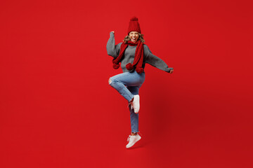 Full body young excited woman wear grey sweater scarf hat jump high do winner gesture isolated on plain red background studio portrait Healthy lifestyle ill sick disease treatment cold season concept