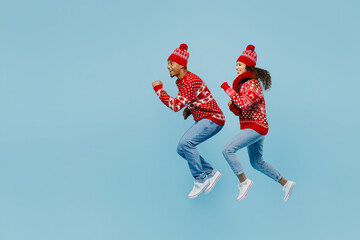 Fototapeta na wymiar Full body side view merry fun young couple two man woman wear red Christmas sweater Santa hat posing jump high run isolated on plain pastel light blue background. Happy New Year 2023 holiday concept.