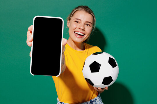 Fototapeta Young woman fan in basic yellow t-shirt cheer up support football sport team hold soccer ball watch tv live stream use mobile cell phone closeup blank screen isolated on dark green background studio