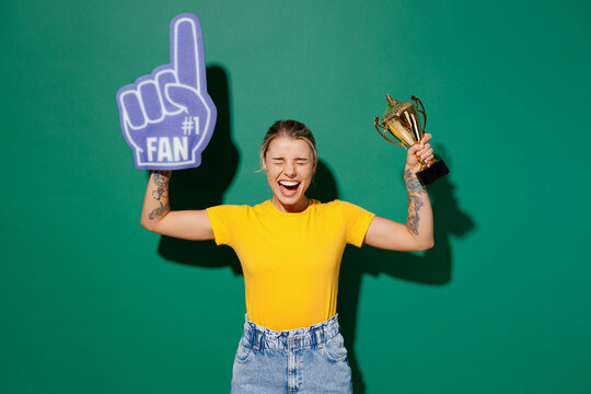 Young woman fan wear basic yellow t-shirt foam 1 glove finger up cheer up support football sport team hold in hand golden cup soccer ball watch tv live stream isolated on dark green background studio.