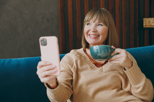 Elderly smiling fun woman 50s year old in casual clothes sit on blue sofa do selfie photo shot mobile cell phone drink coffee stay at home flat rest relax spend free spare time in living room indoor