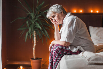 Sad unhappy senior woman seated on bed in bedroom