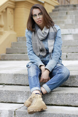 Young girl with dark hair is sitting on the stairs and looking into the frame. She's wearing glasses and jeans