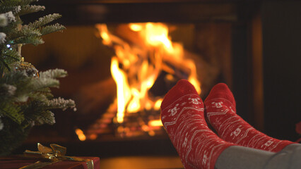 CLOSE UP: Woman warming up her feet in Christmas socks and relaxing by fireplace - obrazy, fototapety, plakaty