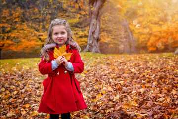 A portrait of a cute, blonde girl in a coat holding a colorful leaf in the park during golden autumn season