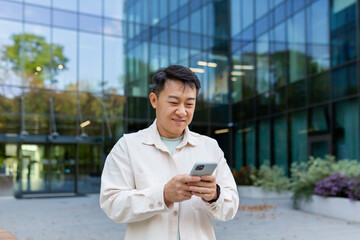 Asian man using phone outside office building, freelancer businessman typing message and smiling, programmer in casual clothes browsing online web pages from app.