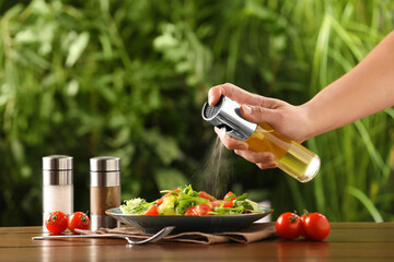 Woman spraying cooking oil onto delicious salad at wooden table against blurred green background,...