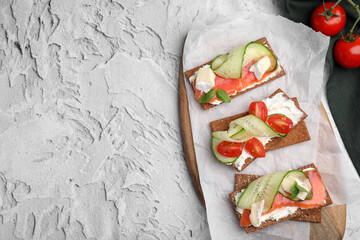 Tasty rye crispbreads with salmon, cream cheese and vegetables on grey textured table, flat lay....