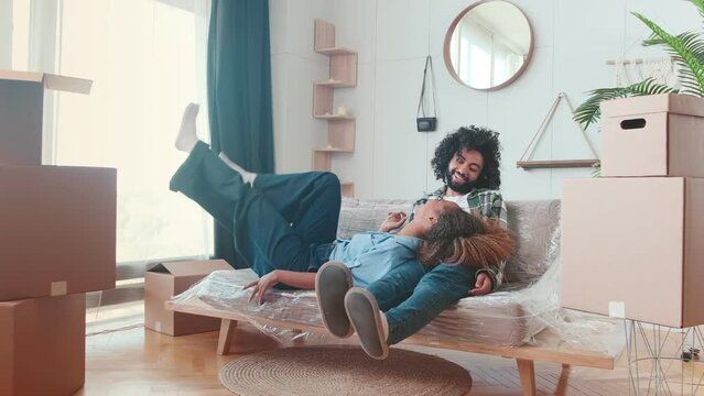 Young inspired happy Arabian man and African American woman fall on sofa and laugh after finishing moving boxes to new apartment bought for young family located in cozy house. Multiracial lifestyle