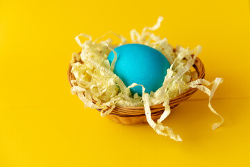 Easter egg painted blue in a basket with artificial straw on yellow background