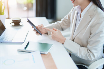 Portrait of a young Asian woman showing a smiling face as she uses her phone, computer and financial documents on her desk in the early morning hours