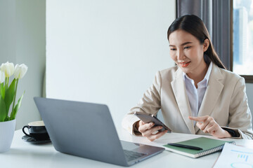 Portrait of a young Asian woman showing a smiling face as she uses her phone, computer and financial documents on her desk in the early morning hours