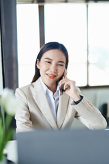 Fototapeta premium Portrait of a young Asian woman showing a smiling face as she uses her phone, computer and financial documents on her desk in the early morning hours